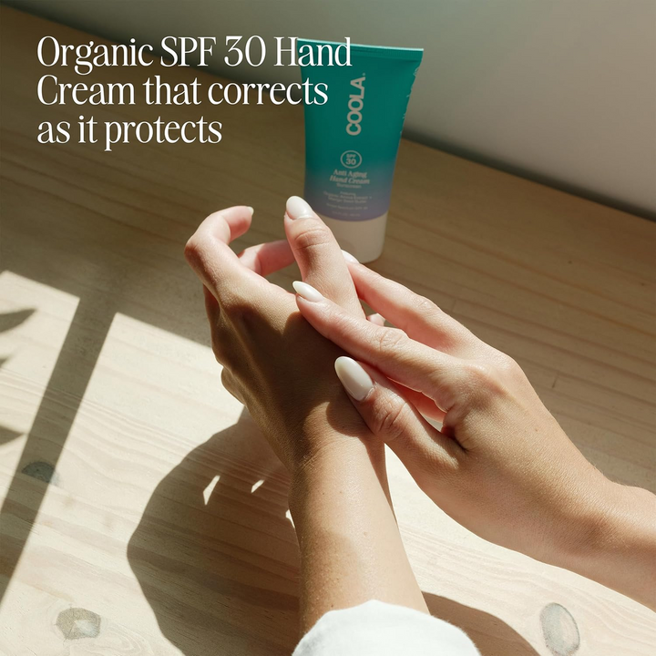Person applying COOLA Organic SPF 30 Hand Cream on hands with a wooden surface in the background.