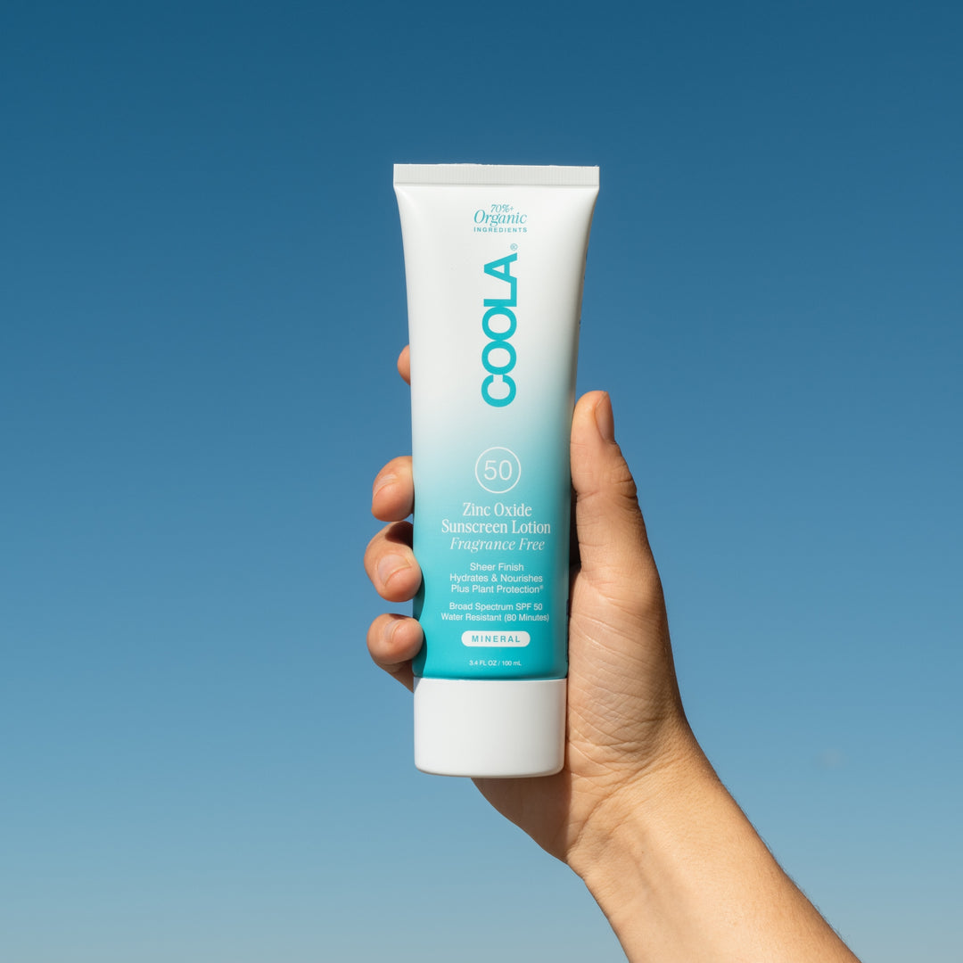 hand holiding COOLA  Mineral Body Organic Sunscreen Lotion SPF 50 - Fragrance Free at the beach