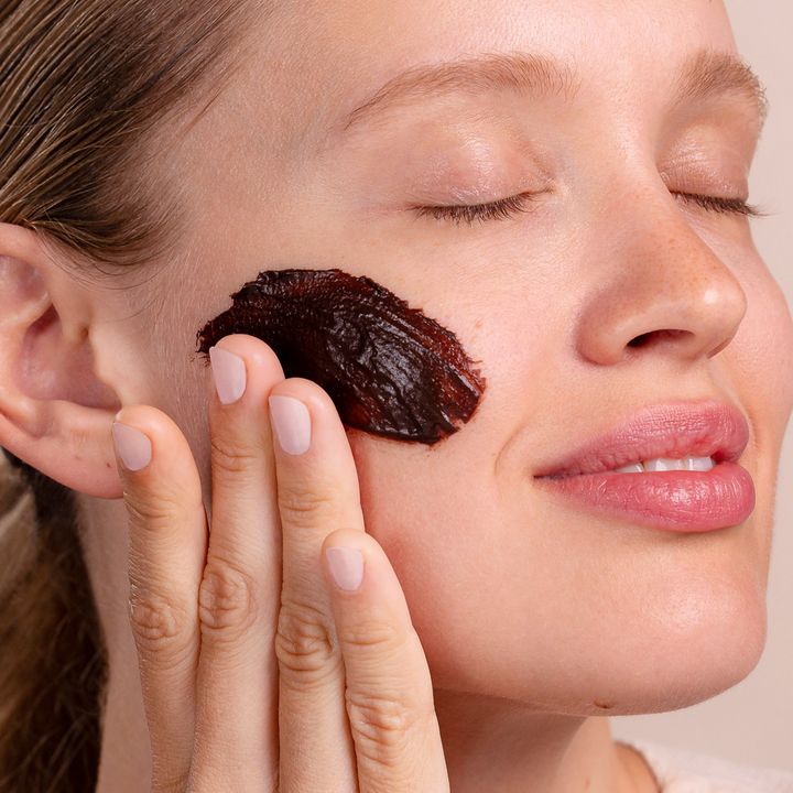Woman applying Eminence Organics Chocolate Mousse Hydration Masque to her cheek with a neutral background