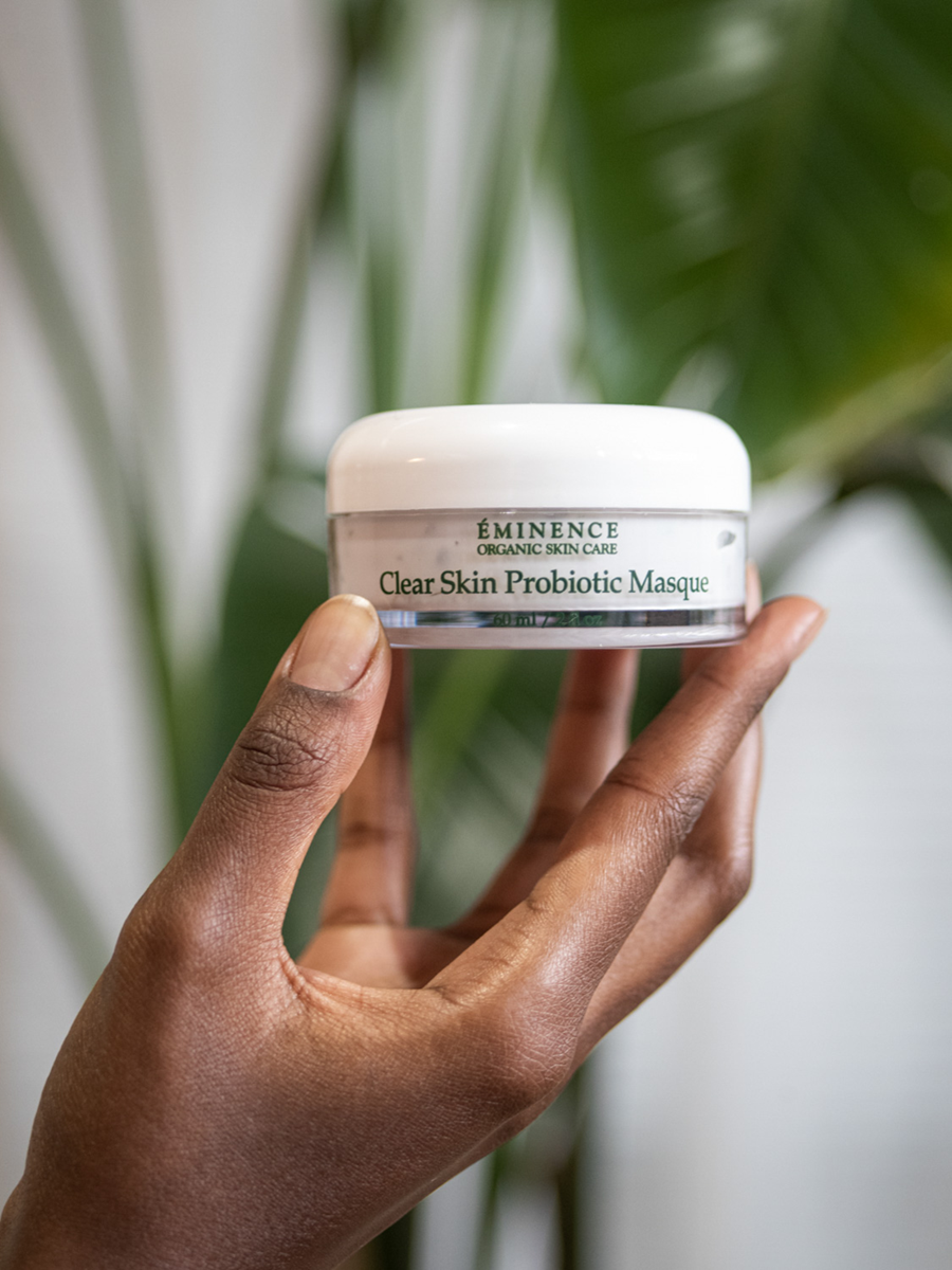 Hand holding a jar of Eminence Clear Skin Probiotic Masque against a blurred green leafy background