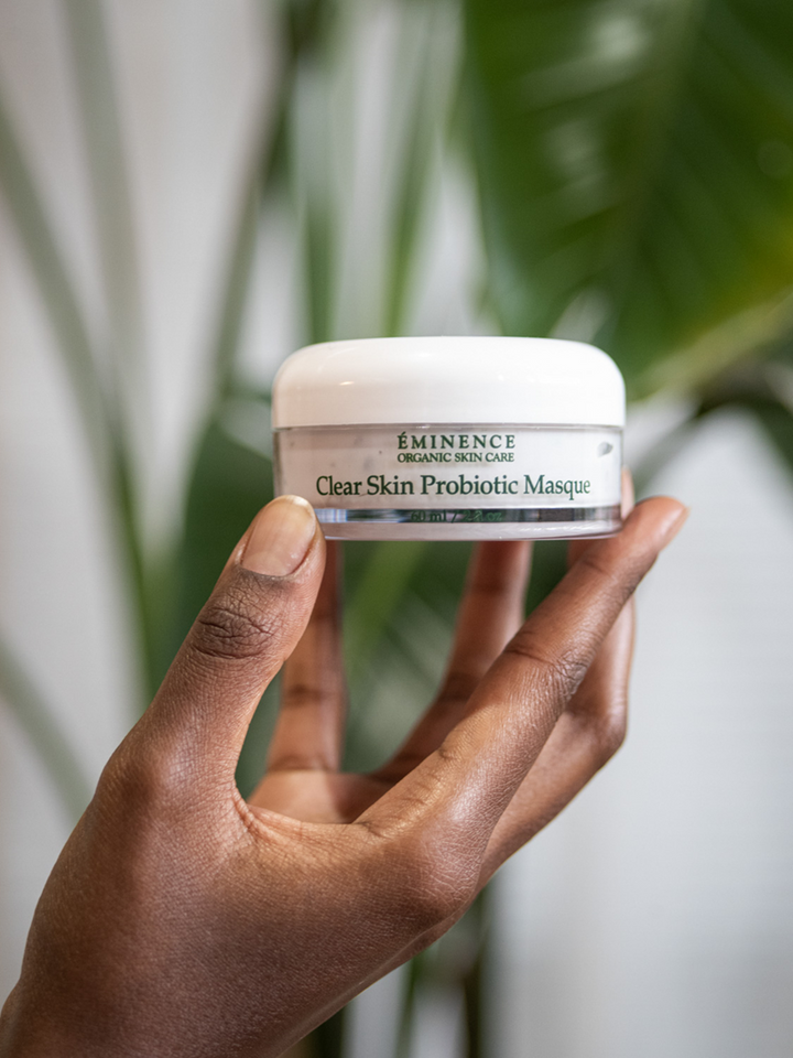 Hand holding a jar of Eminence Clear Skin Probiotic Masque against a blurred green leafy background