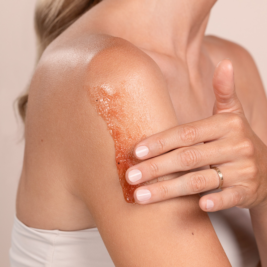 Person applying Eminence Organics Cranberry Pomegranate Sugar Scrub to their shoulder with a neutral background