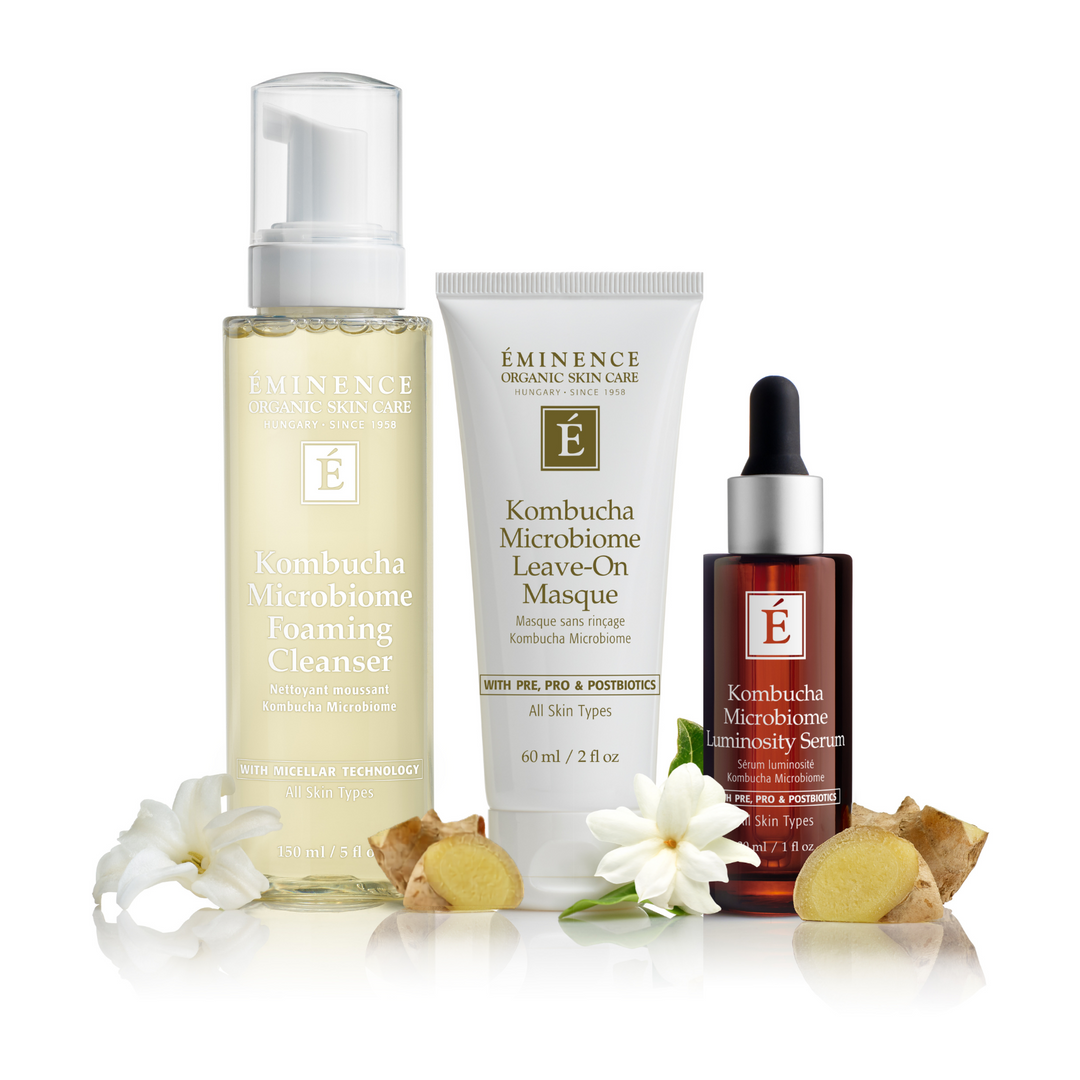 Three skincare products from Eminence Organic Skin Care arranged on a white background with flowers and ginger slices: a foaming cleanser bottle, a leave-on masque tube, and a microsyser serum dropper bottle.