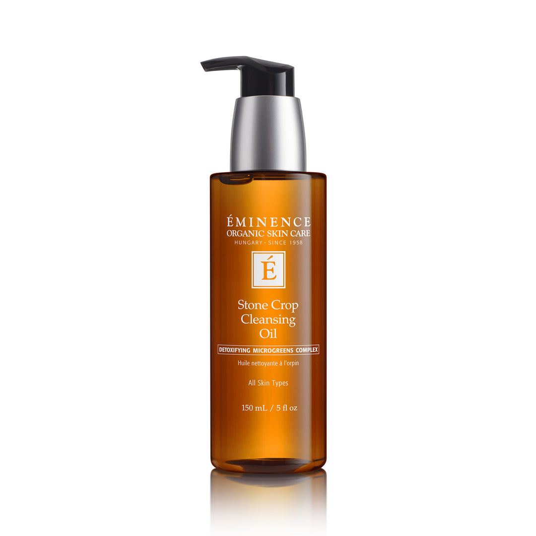 A bottle of Éminence Organic Skin Care Stone Crop Cleansing Oil on a white background with a soft reflection. The bottle is amber brown with a black and silver pump dispenser.