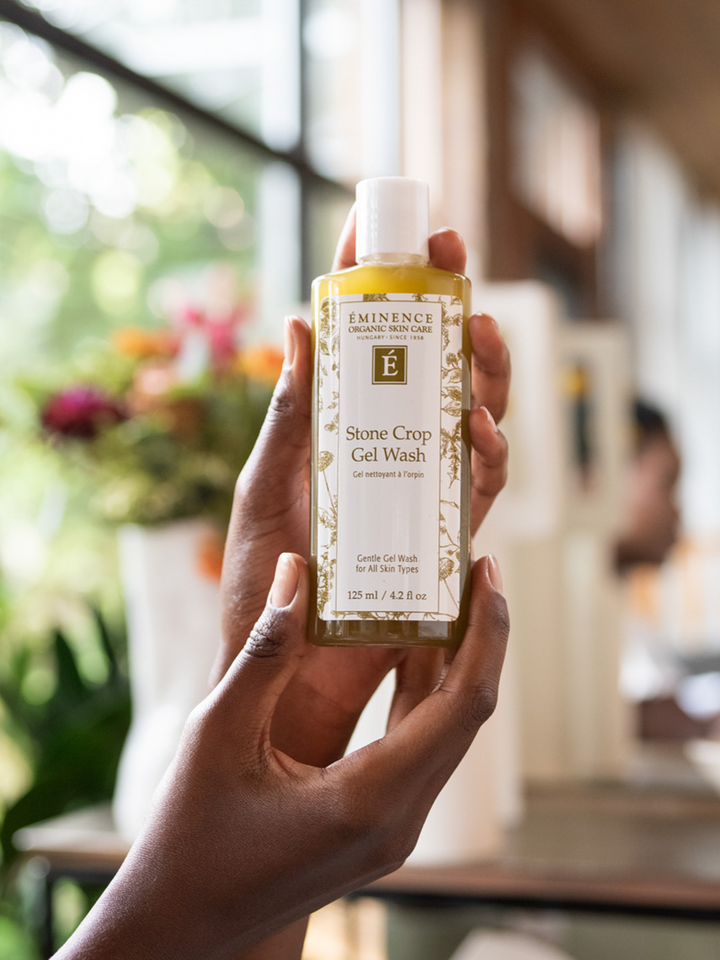 Hand holding a bottle of Eminence Stone Crop Gel Wash in a blurred indoor setting