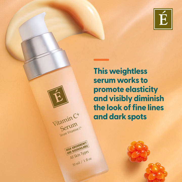A bottle of Eminence Organics Vitamin C+ Serum with a silver pump and frosted bottle, set against a peach-colored background with a cream-colored serum smear, French text describing the product benefits, and small orange berry illustrations.