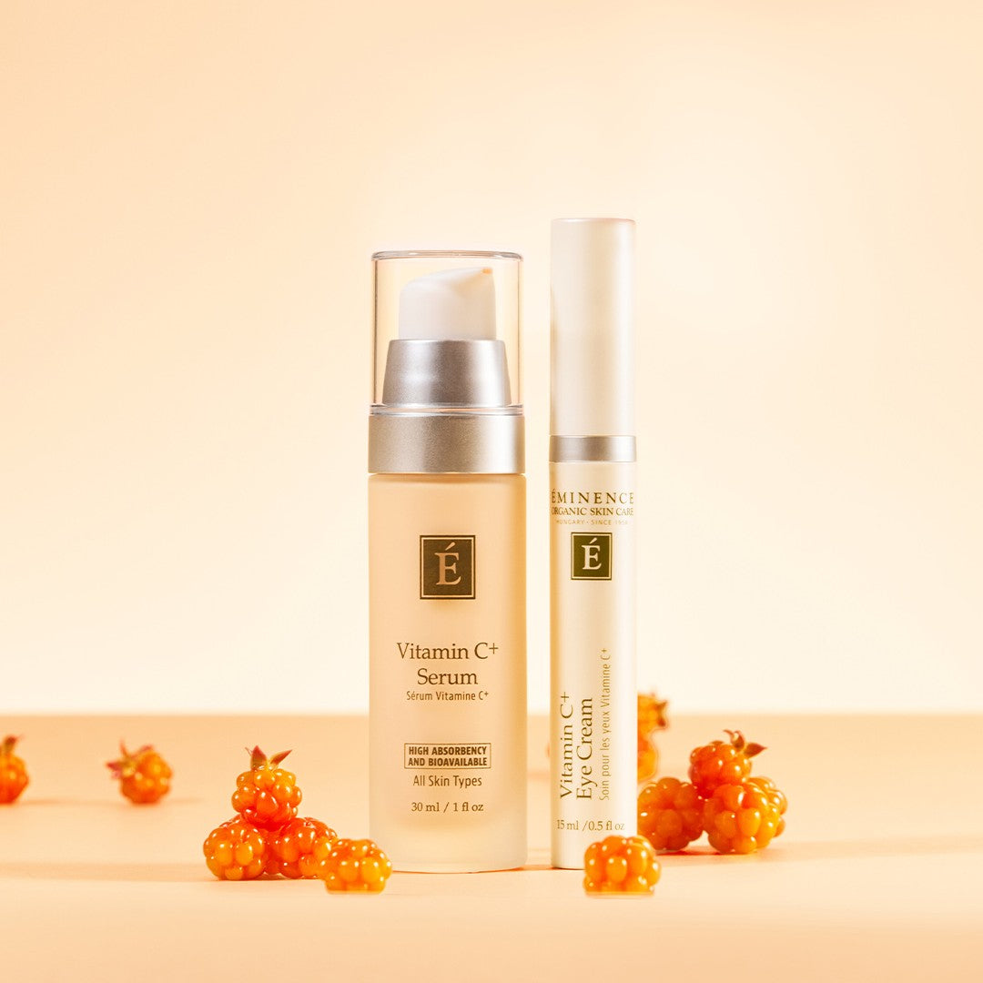 A skincare set featuring a beige Eminence Organics Vitamin C Serum bottle and Eminence Organic Vitamin C Eye Cream tube styled with small orange berries on a warm beige background.