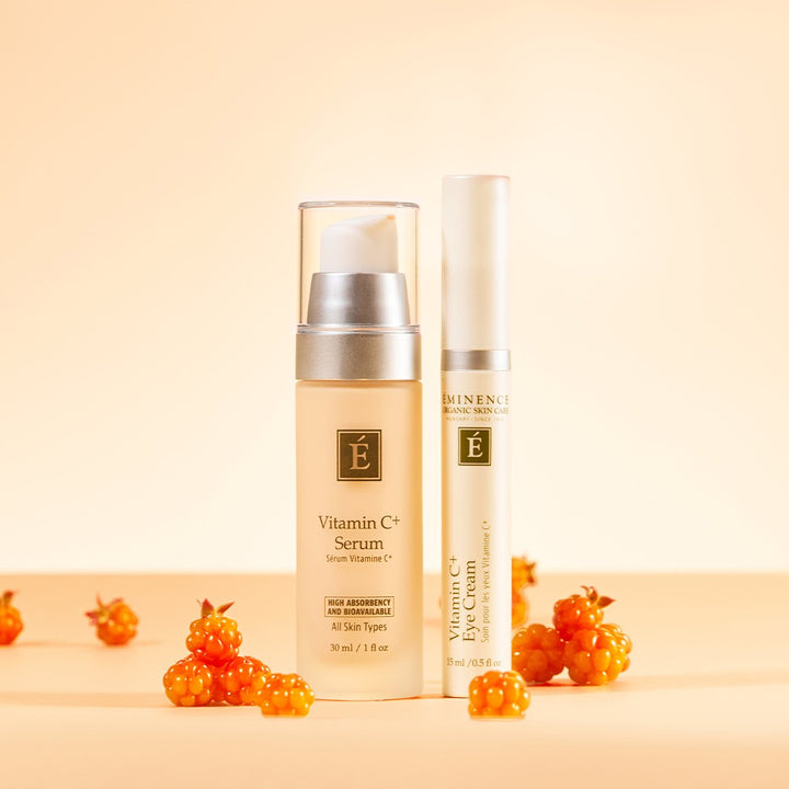 A skincare set featuring a beige Eminence Organics Vitamin C Serum bottle and Eminence Organic Vitamin C Eye Cream tube styled with small orange berries on a warm beige background.