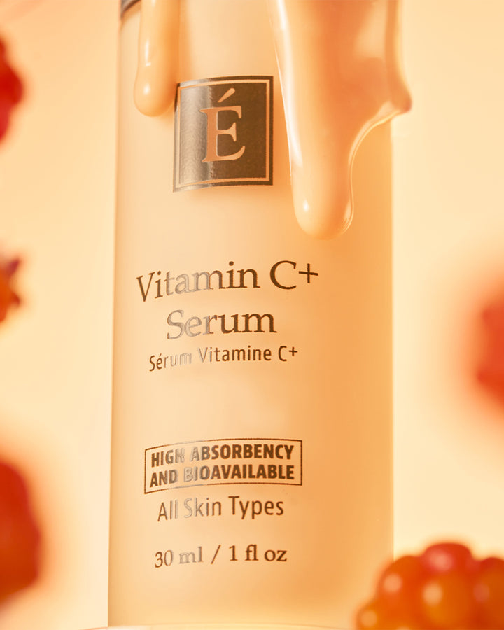A close-up of Eminence Organics Vitamin C+ Serum with a warm peachy-orange glow and glossy cream dripping from the top. The bottle is centered against a softly blurred background with small red fruit-like shapes at the edges.