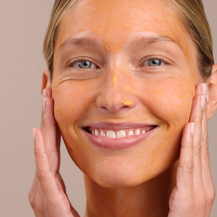 Woman applying Eminence Organics Yam &amp; Pumpkin Enzyme Peel 5% to her face with a neutral background