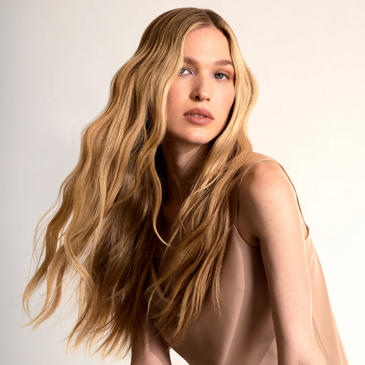 A woman with long, wavy blonde hair wearing a sheer beige satin top, photographed against a clean off-white studio background.