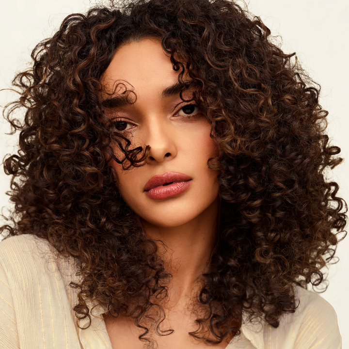 A close-up portrait of a woman with voluminous, tightly curled brown hair, wearing a cream-colored top against a clean, light off-white background.