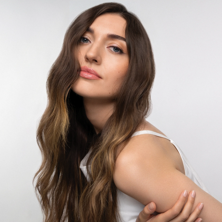 Model with long, wavy brown hair resting a hand on her upper arm against a light background