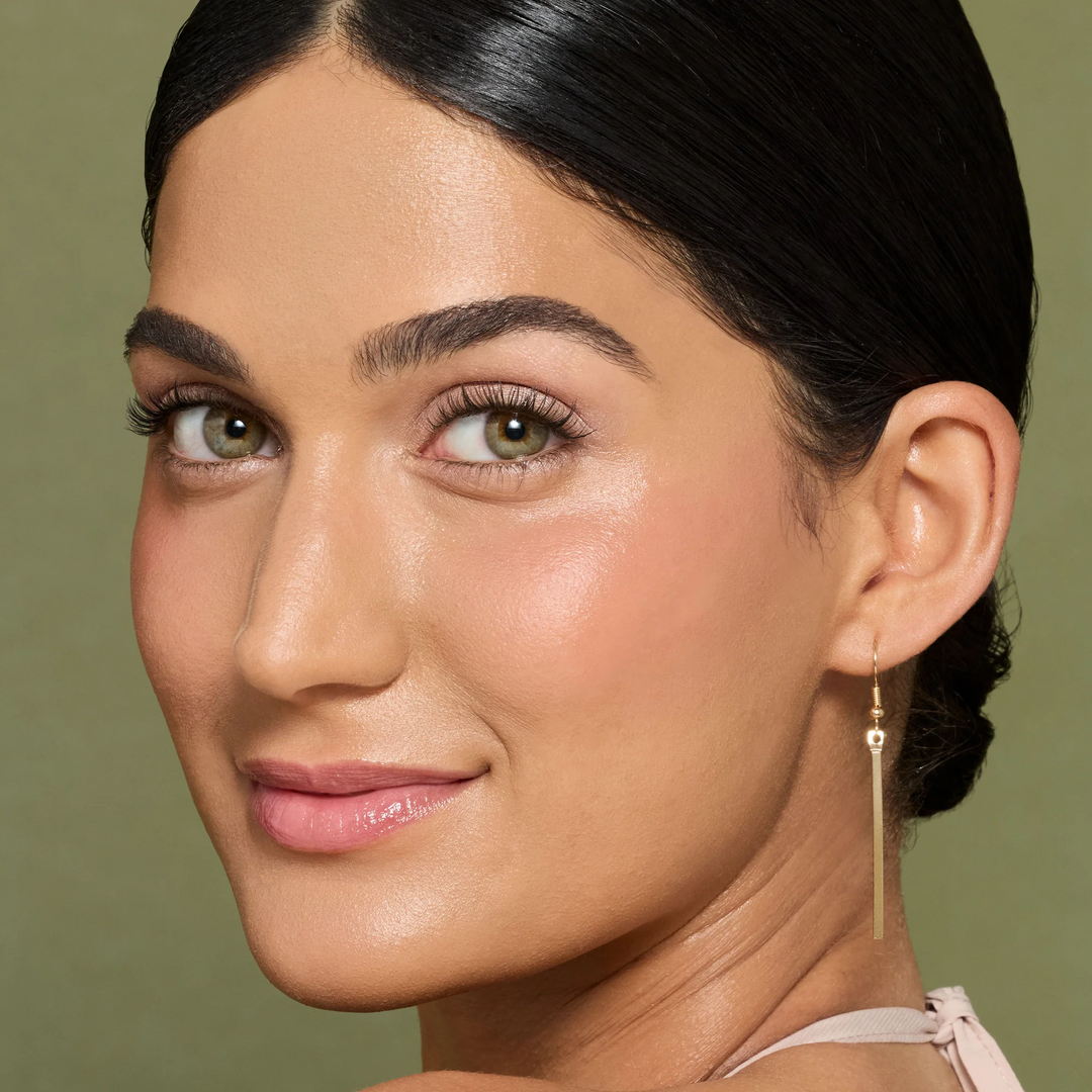 Close-up of a dark-haired model with shimmery pink eye shadow and defined lashes against an olive-green background.
