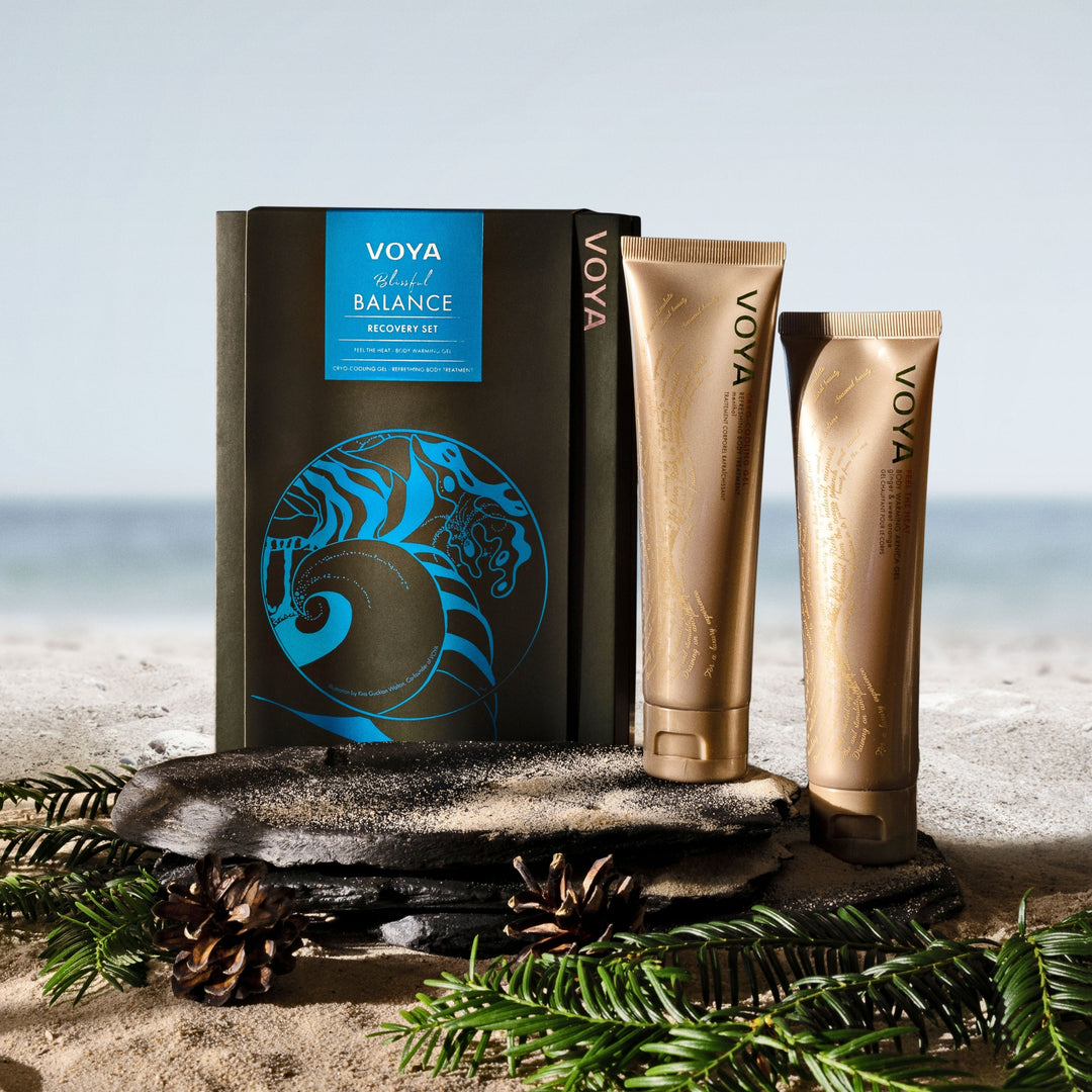 VOYA Blissful Balance Recovery Set displayed on slate and sand beside the sea, with evergreen branches and pine cones.
