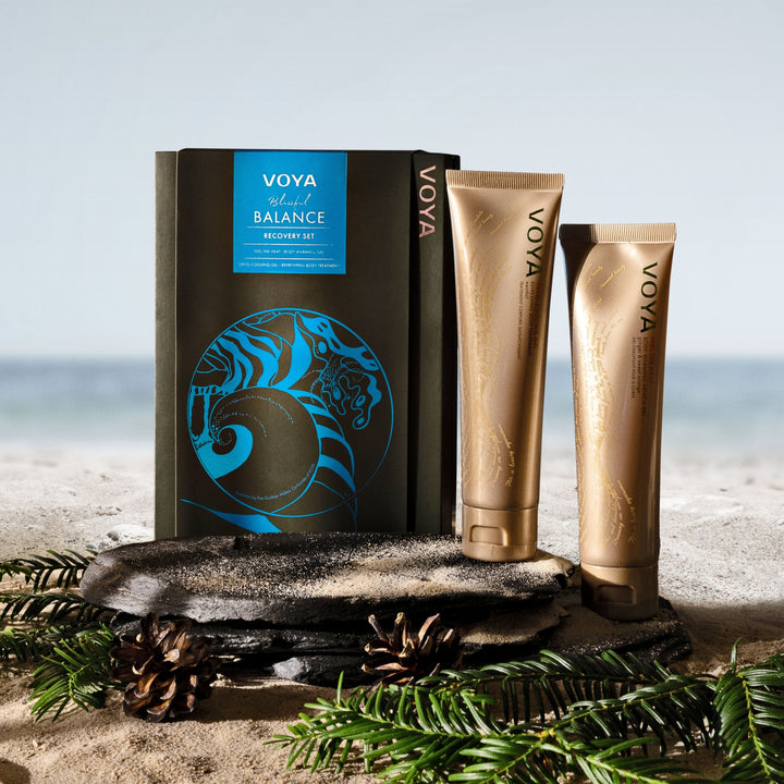 VOYA Blissful Balance Recovery Set displayed on slate and sand beside the sea, with evergreen branches and pine cones.
