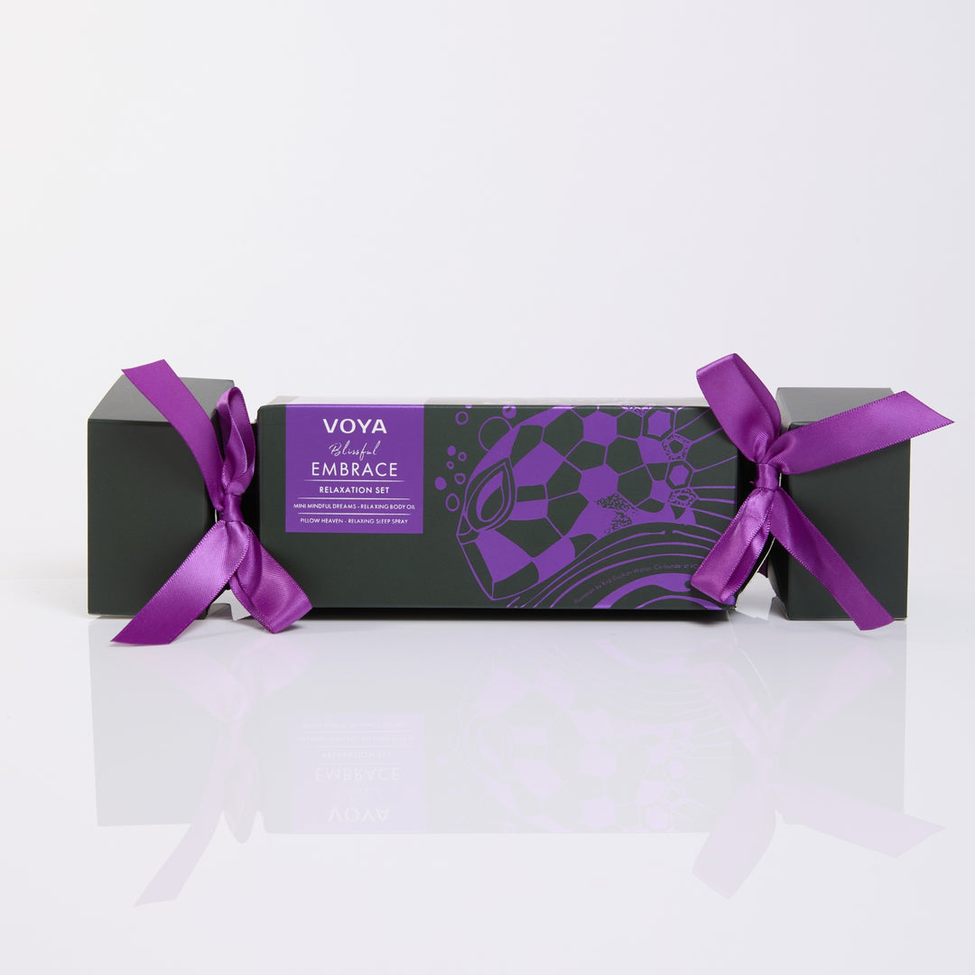 VOYA Blissful Embrace cracker-shaped gift box with purple ribbon bows and purple illustrations on dark green packaging.