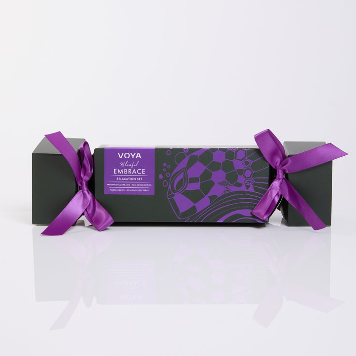 VOYA Blissful Embrace cracker-shaped gift box with purple ribbon bows and purple illustrations on dark green packaging.