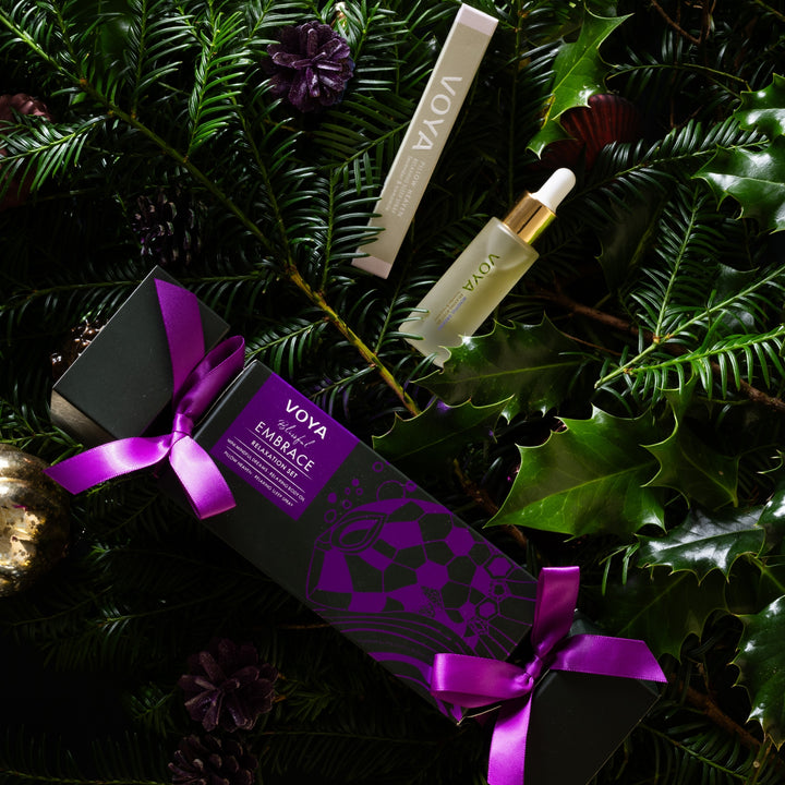 VOYA Blissful Embrace gift set nestled among evergreen branches, holly leaves and festive ornaments.
