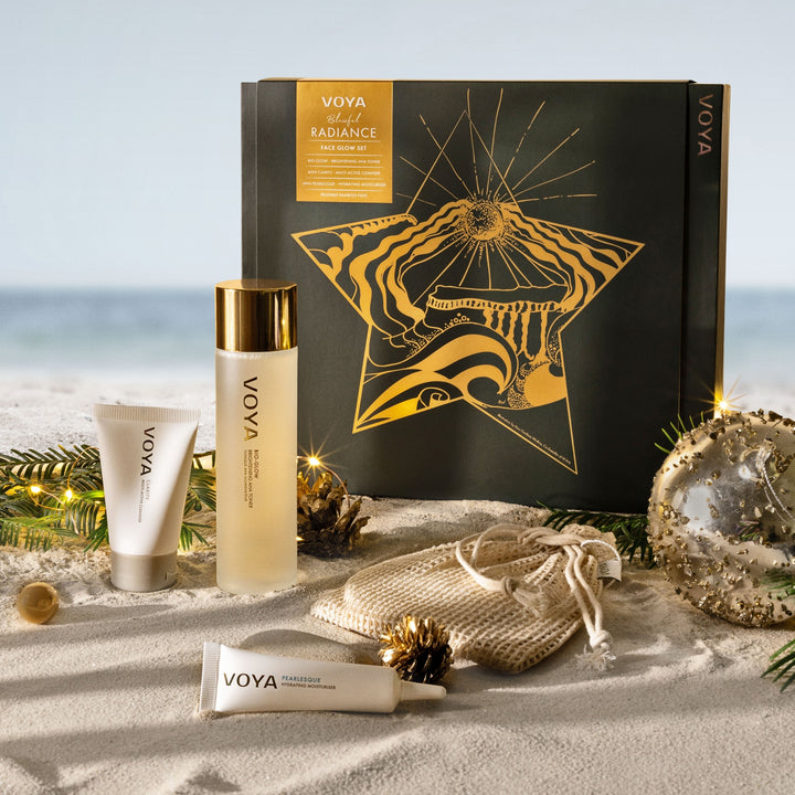 VOYA Blissful Radiance Face Glow Set displayed on sand with festive greenery, string lights and gold ornaments.
