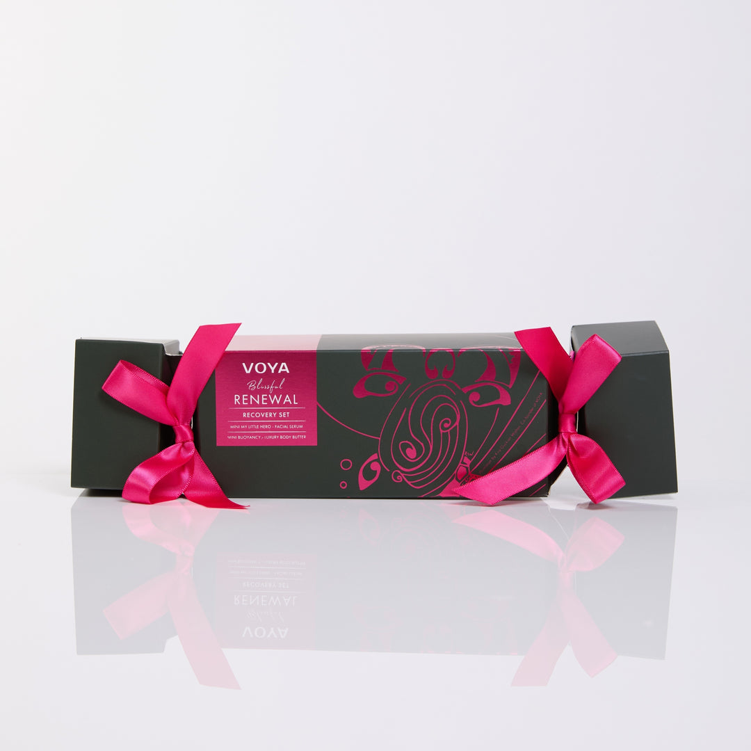 VOYA Blissful Renewal cracker-shaped gift box with pink ribbon bows and pink illustrations on dark green packaging.