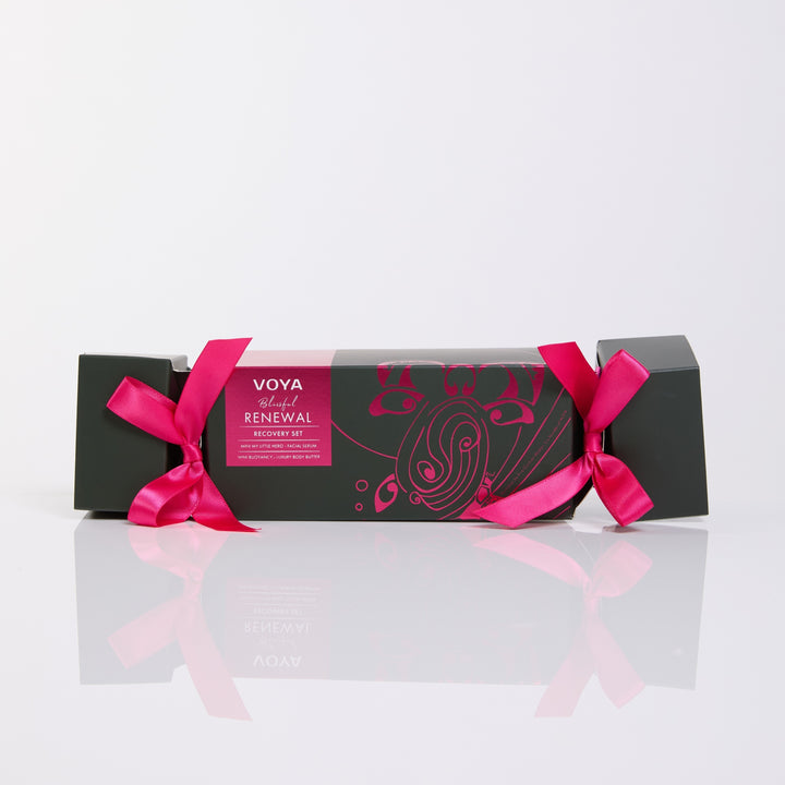 VOYA Blissful Renewal cracker-shaped gift box with pink ribbon bows and pink illustrations on dark green packaging.