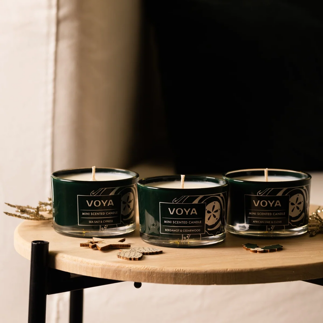 Three VOYA mini candles arranged on a round wooden table with small festive ornaments.

