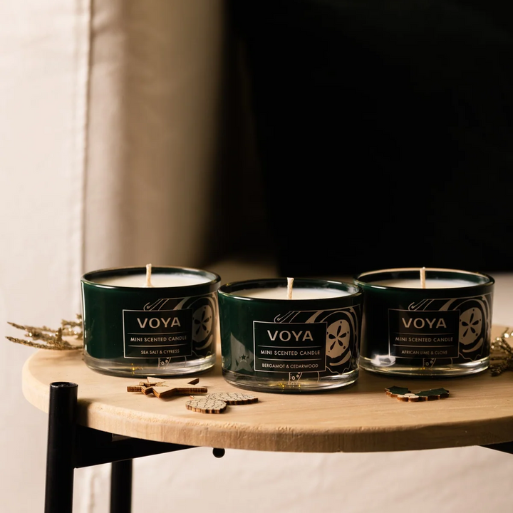 Three VOYA mini candles arranged on a round wooden table with small festive ornaments.
