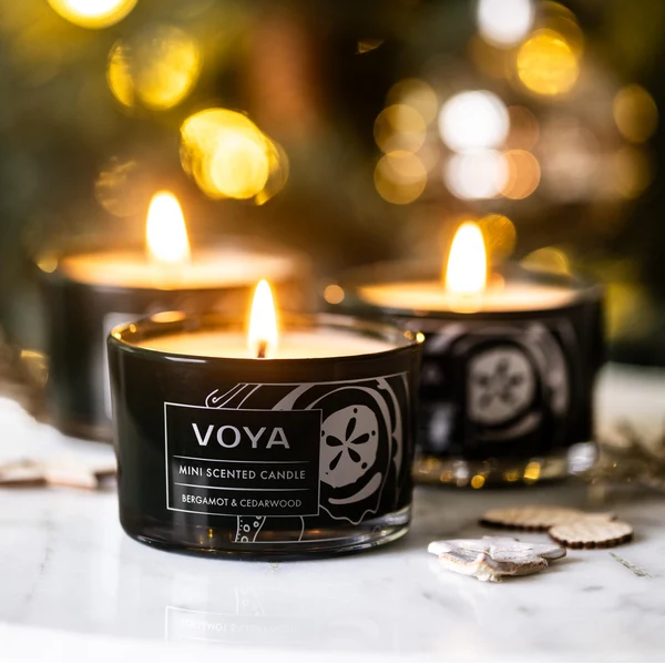VOYA Blissful Sparks mini candles glowing beside festive decorations, with Bergamot &amp; Cedarwood in the foreground.

