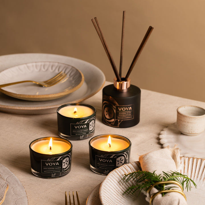 Three lit VOYA mini candles on a festive dining table beside a reed diffuser and gold-trimmed tableware.
