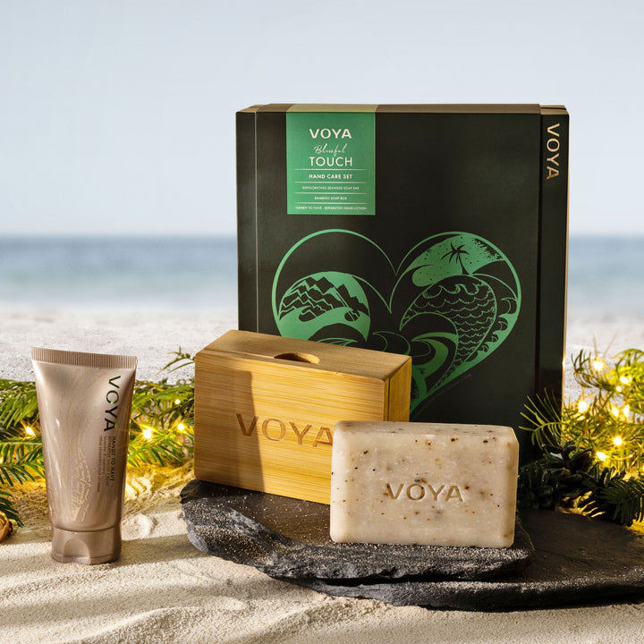 VOYA Blissful Touch Hand Care Set displayed on sand beside the sea with festive greenery and string lights.
