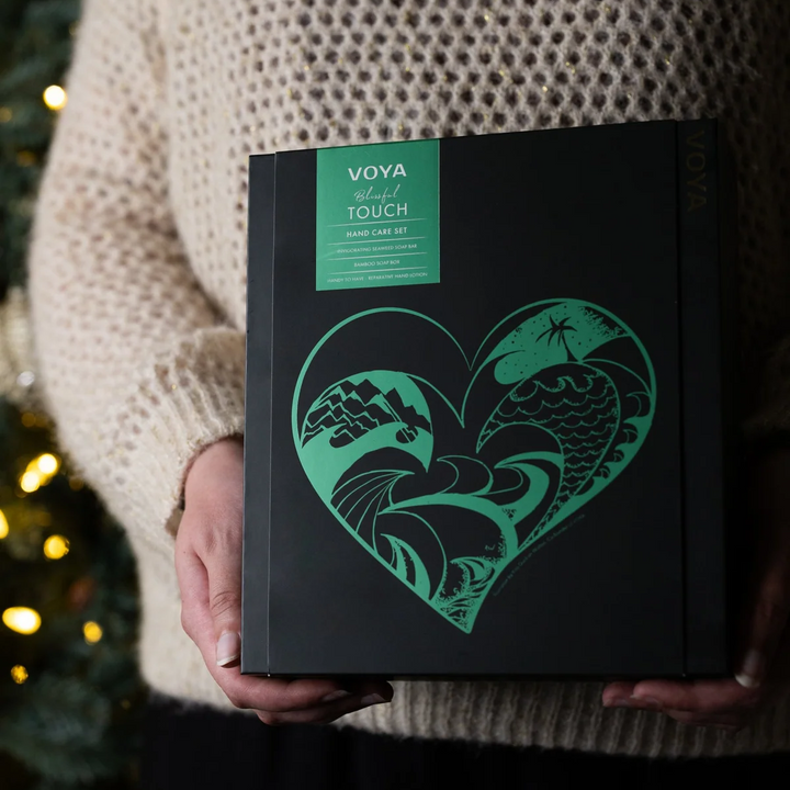 Person in a cream knit sweater holding the VOYA Blissful Touch gift box with a green heart-shaped coastal illustration.
