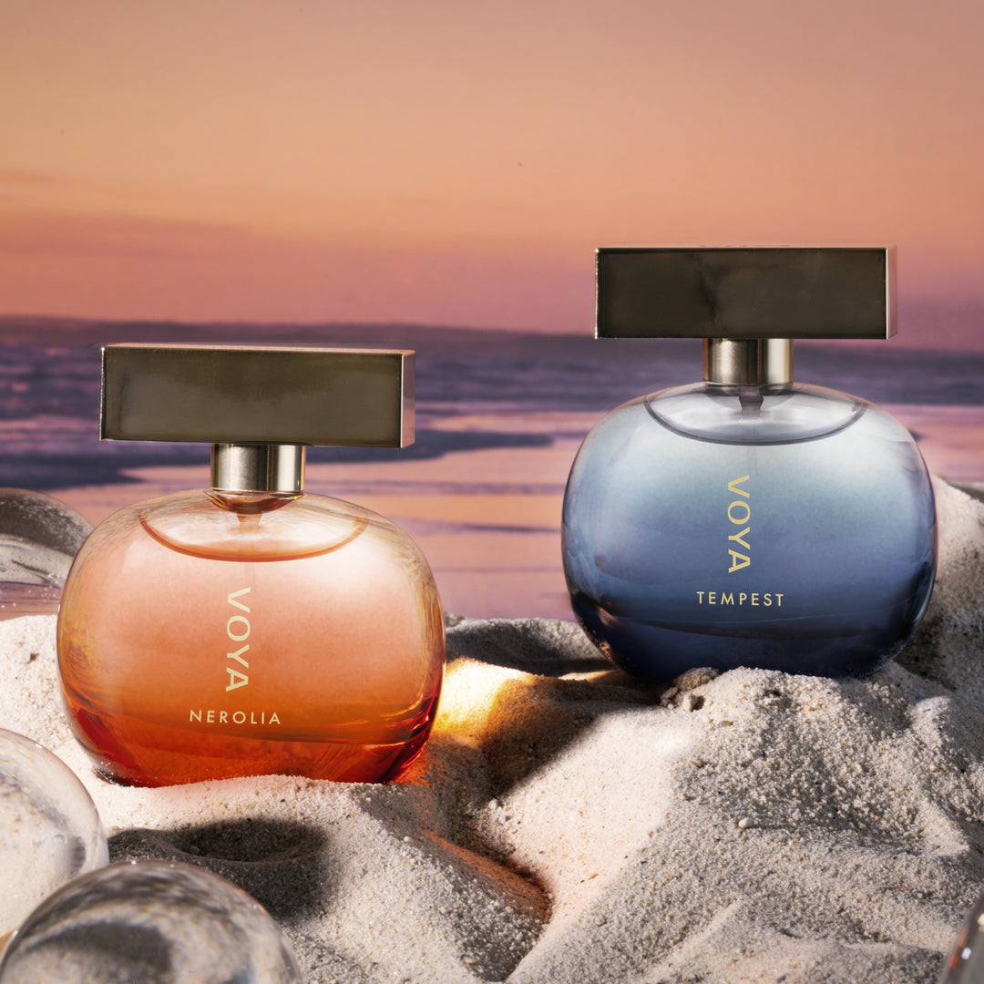 Two Voya perfume bottles, Nerolia in a peachy-orange shade and Tempest in a blue-gray shade, displayed on white sand with ice cubes against a sunset beach background.