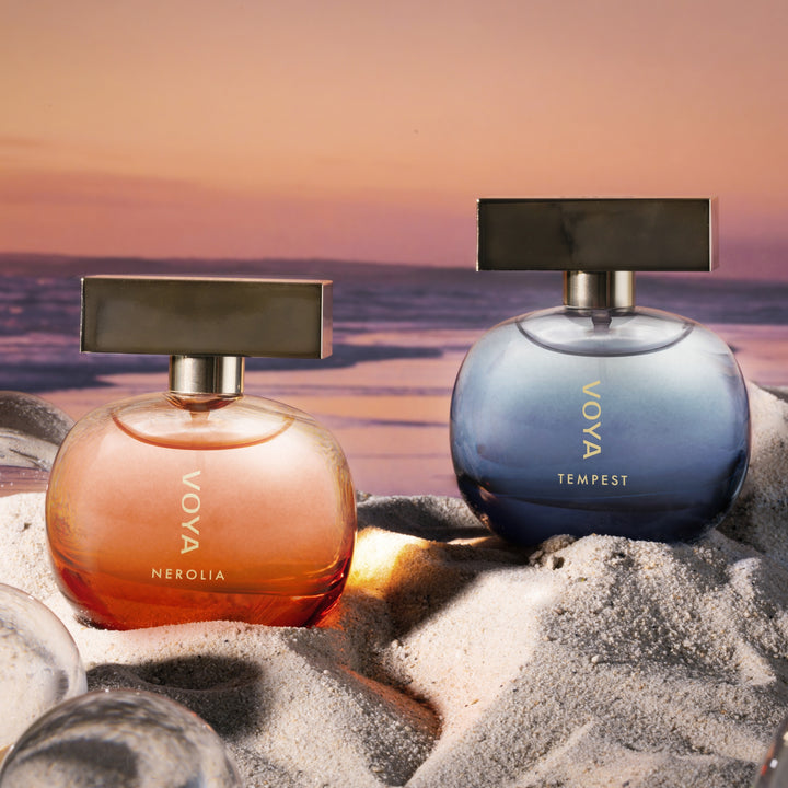 Two Voya perfume bottles, Nerolia in a peachy-orange shade and Tempest in a blue-gray shade, displayed on white sand with ice cubes against a sunset beach background.