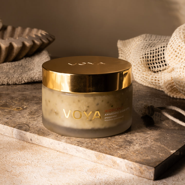VOYA Iridescence Awakening Sugar Scrub on a stone tray beside a body brush and mesh bag