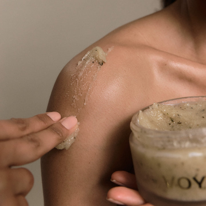 Person applying VOYA Iridescence Awakening Sugar Scrub to their shoulder while holding the open jar