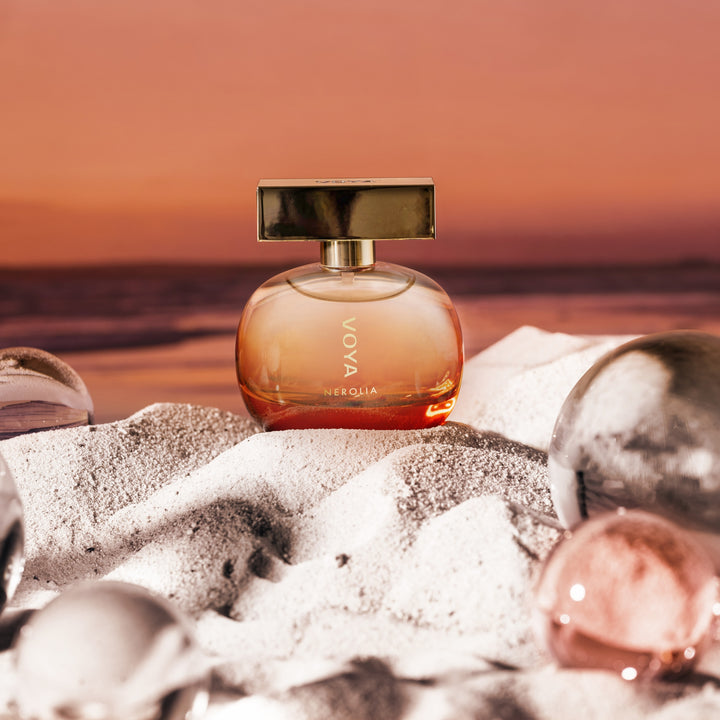 A perfume bottle labeled VOYA Nerolia - Eau de Toilette sits on white sand with reflective stones, set against a warm sunset beach background.