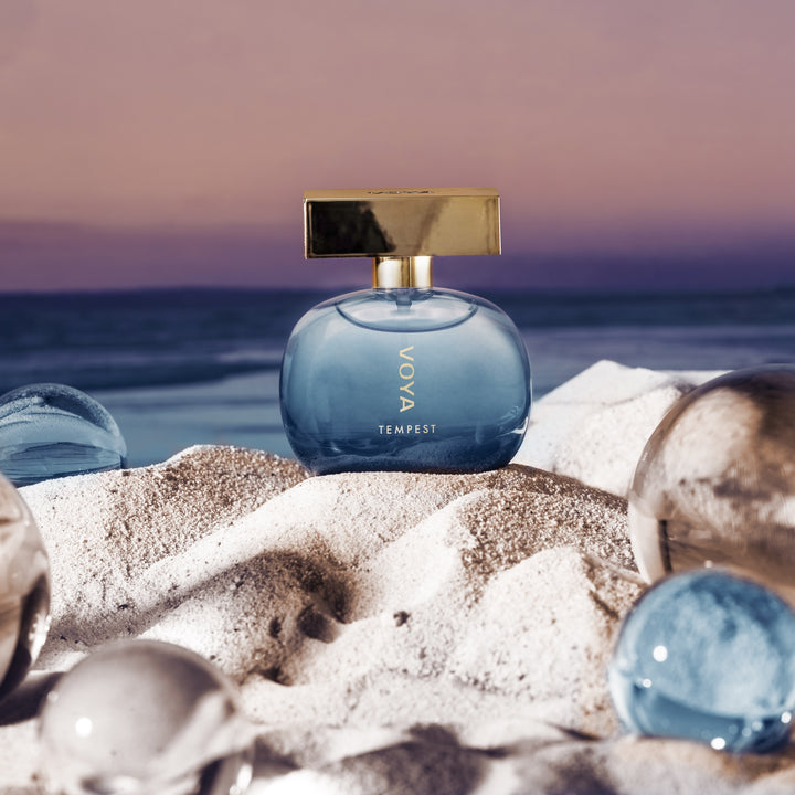 A round blue glass perfume bottle with a gold cap sits on white sand with glass marbles in the foreground and a beach sunset in the background. The bottle is labeled &