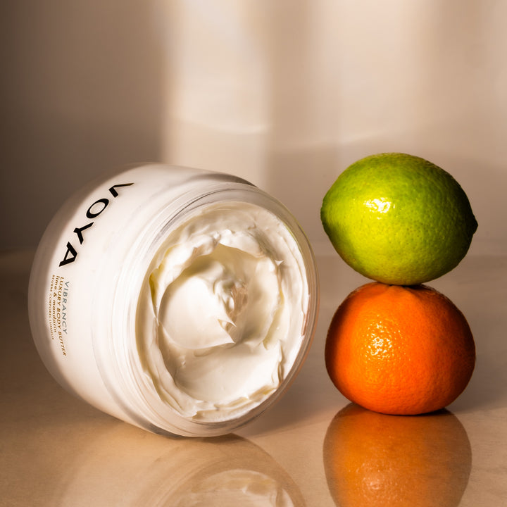 Open jar of VOYA Vibrancy Luxury Body Butter showing its creamy texture beside a lime and mandarin.
