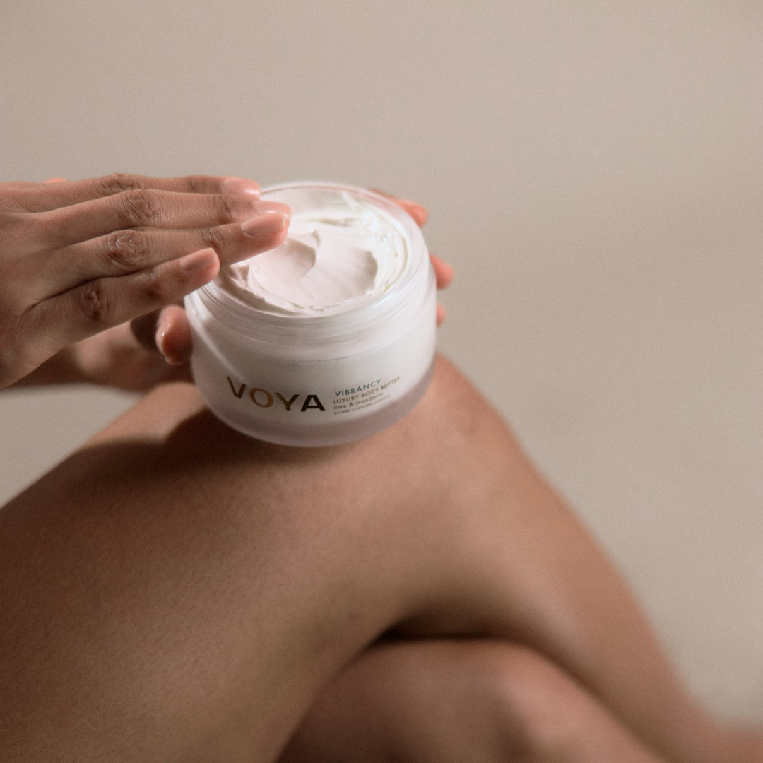 Person scooping VOYA Vibrancy Luxury Body Butter from an open jar resting on their knee.

