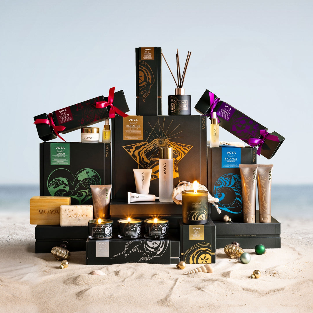 VOYA holiday collection featuring gift sets, skincare, candles and a reed diffuser arranged on sand with festive ornaments.