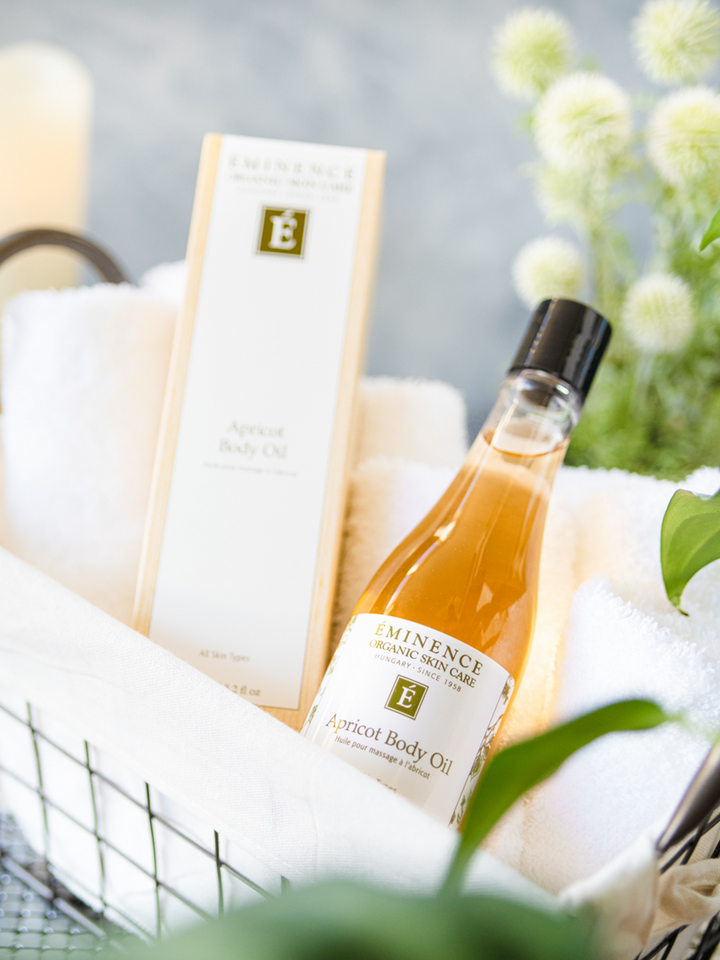 Bottle of Eminence apricot body oil in a basket with a towel and flowers in the background