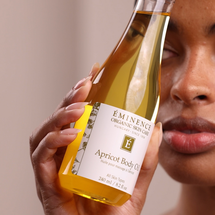 Hand holding a bottle of Eminence Organic Skin Care Apricot Body Oil with a blurred face in the background.