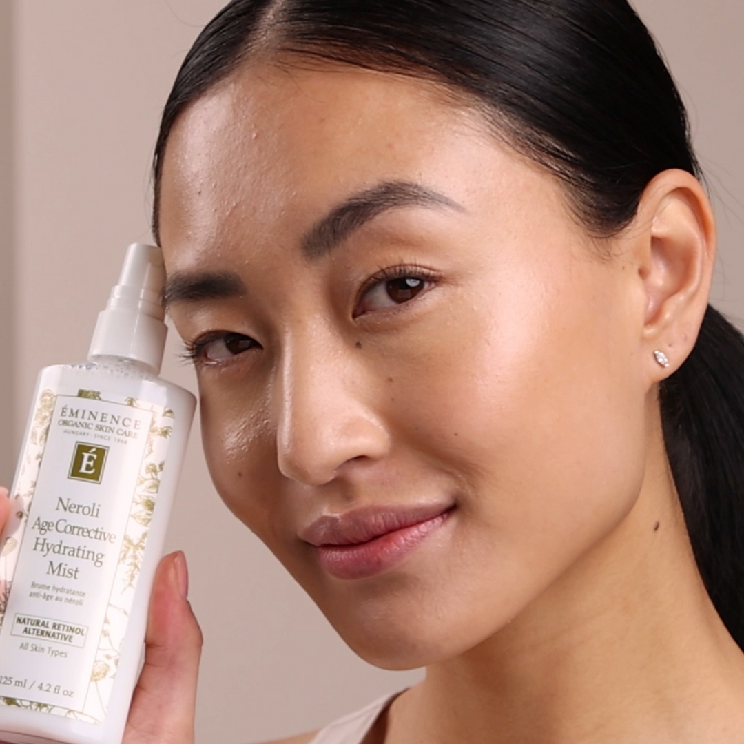 Woman holding Eminence Organics Neroli Age Corrective Hydrating Mist near her face against a neutral background